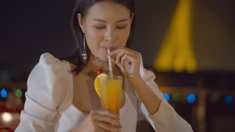 stunning asian woman with an orange cocktail at a rooftop bar and the city, bokeh lights in the background