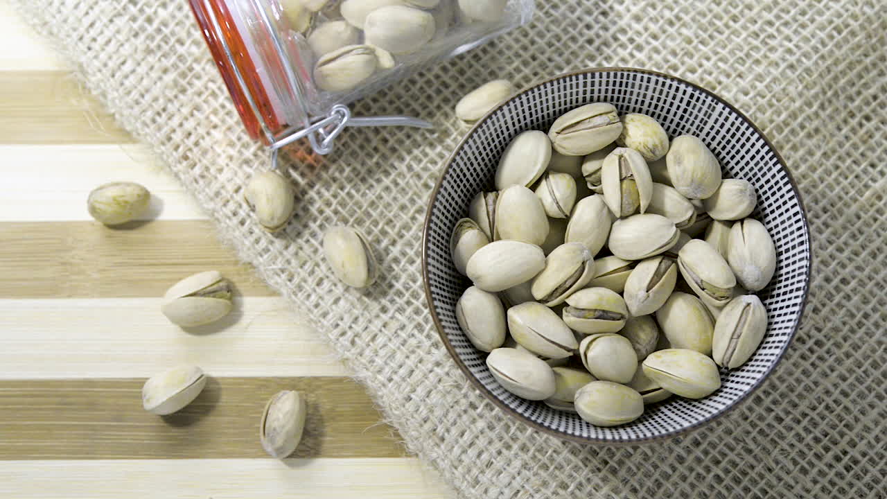 toma panorámica lenta vista de arriba hacia abajo sobre un tazón de cerámica de un frasco de vidrio lleno de una mezcla de castañas con pasas sobre una mesa a rayas con tela de arpillera