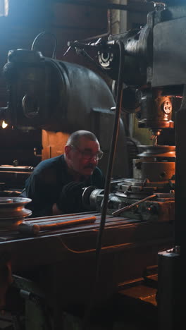 mature worker with glasses looks at drilling machine cutting out holes to control operation in factory workshop. lathe equipment processes steel detail. vintage plant