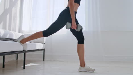 brunette in black sportswear in a white apartment makes a split squat with dumbbells in her hands leaning on the sofa with her foot.