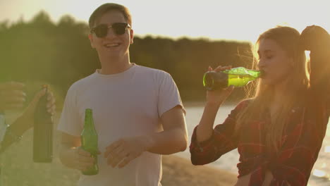 loving couple celebrate the end of the semester with beer and pop music on the beach with their friends. girl is dancing on the open air party and drinking beer at sunset in hot summer evening.