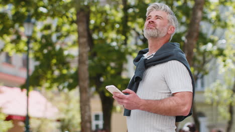 Tourist-mature-man-using-smartphone-text-messages-looking-for-search-a-way-on-map-in-navigator-app