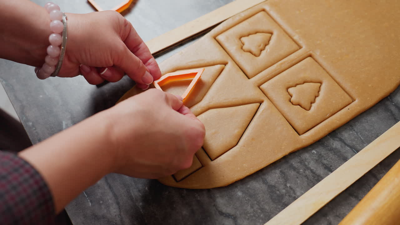 vista de la mano de la mujer colocando un cortador en forma de casa naranja en la masa para crear formas de corte precisas, los cortes cuadrados y triangulares son visibles, cuidadosamente dispuestos en la masa marrón lisa para hornear