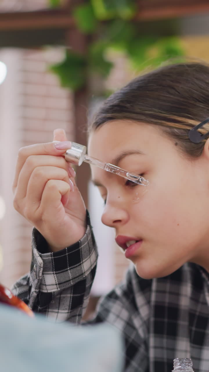 Adolescente aplicándose sérum con cuentagotas en una mejilla con una camisa de cuadros frente al espejo, con cuidado, luz natural de cerca con plantas de interior de fondo, expresión concentrada, ambiente de tutorial, brillo sutil