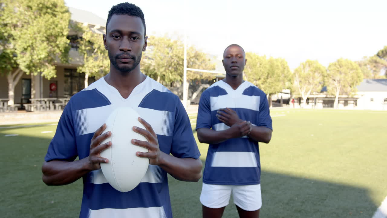 Holding rugby ball, african american male athlete standing on field with teammate, copy space