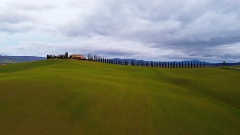 experience an aerial dolly over cinigiano, tuscany, italy's rolling hills. the camera moves forward, capturing a picturesque farmhouse, cypress trees, and lush green fields under a dramatic cloudy sky