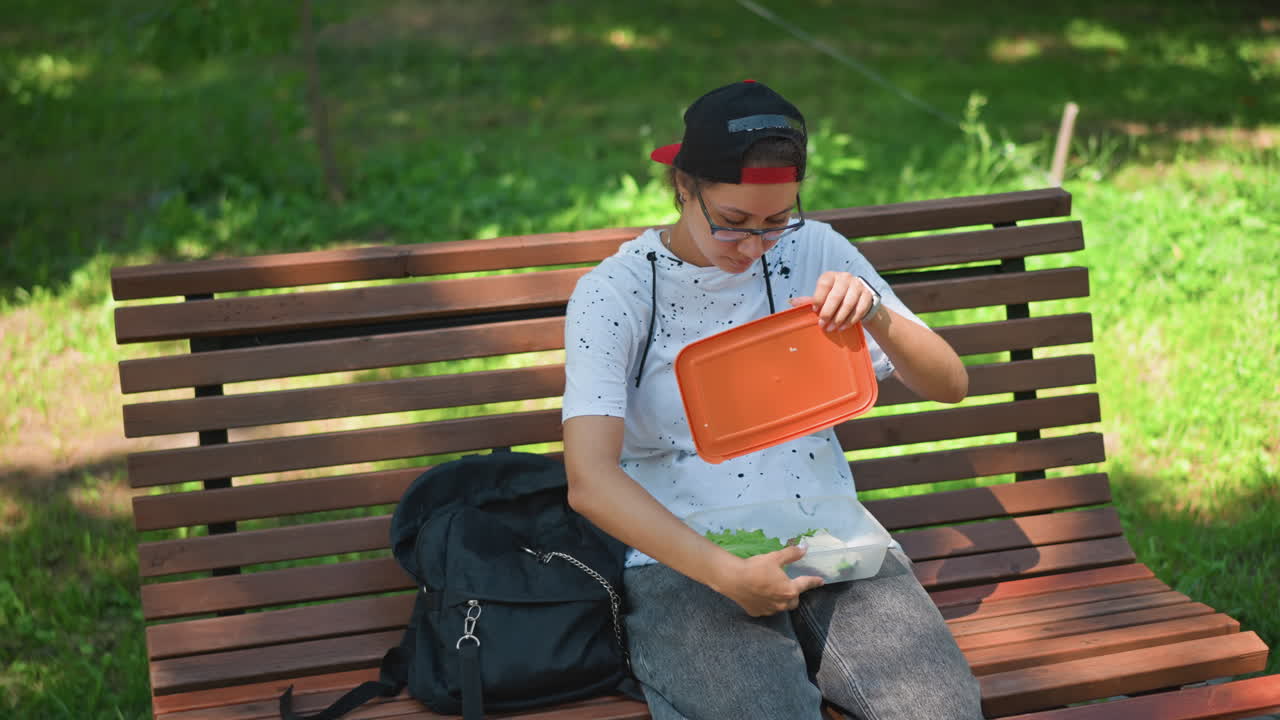 학생이 벤치에서 점심을 꺼내고 있어요. 흰색 점박이 셔츠, 빨간색 모자, 백팩을 메고 나무 아래에서 주황색 파우치를 열어 햇빛이 얼룩덜룩하게 비치는 한가로운 한낮에 가방에 손을 뻗어 샐러드를 먹고 있으며, 이어폰을 끼고 도시의 평온함을 즐기고 있어요.