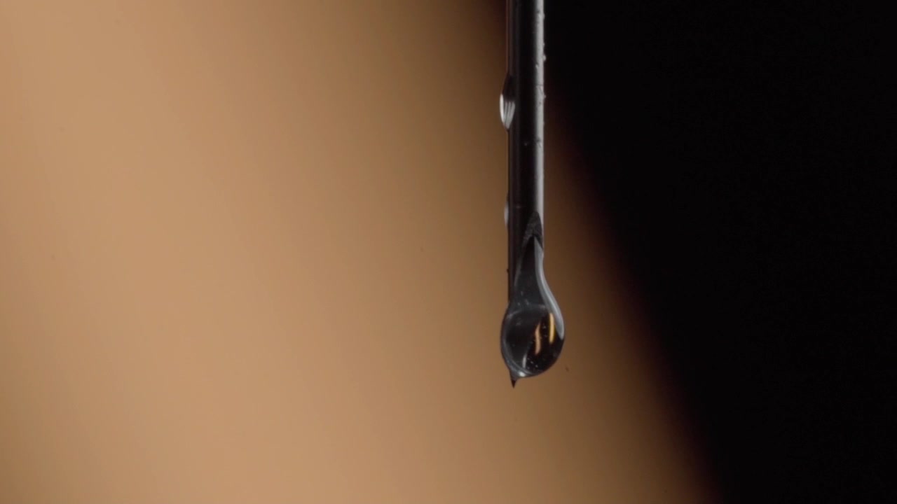 Makronærbilde av en vanndråpe som drypper fra en mørk pinne mot en uskarp bakgrunn. Klar væske dannes på tuppen før den faller, noe som fremhever overflatespenning og glatte reflekterende detaljer.