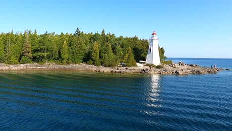 Orbit-Shot-Of-White-Lighthouse-In-Front-Of-Green-Trees-At-Tobermory-Huron-Lake-,-Canada