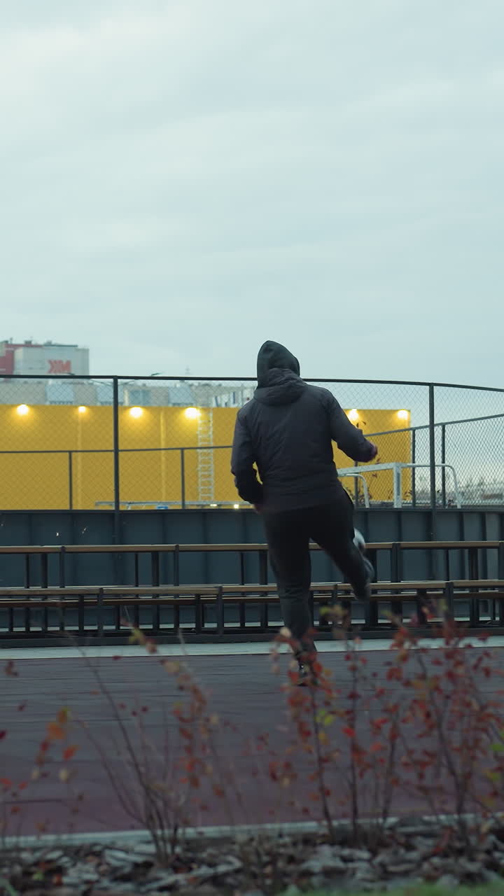 Athlete skillfully juggling football on sport arena surrounded by urban outdoor elements, background includes benches, playground, lighting poles, and urban architecture