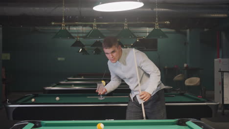 billiards player in white shirt and grey trousers holds cue stick and glass of lemon drink while pacing around pool hall. overhead lights cast a warm ambiance over green pool tables