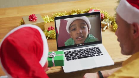 padre caucásico con hijo con sombreros de santa usando una computadora portátil para una videollamada de navidad, con el niño en la pantalla