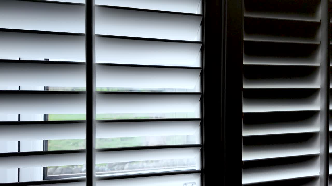 White louvered shutters partially closed, filtering light from a window. close up, dolly right