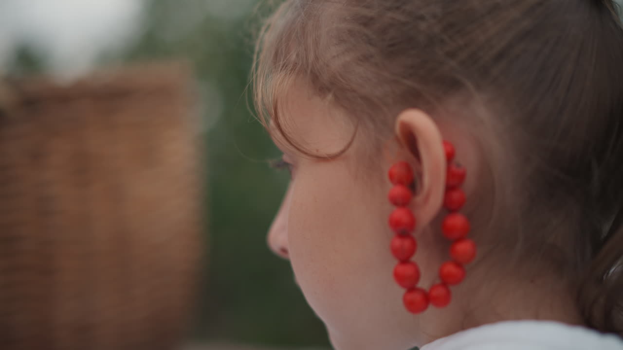 Primo piano di profilo di una ragazza con perline rosse che indossa un grande orecchino di perline vicino a un cesto di vimini, luce morbida esterna, profondità di campo ridotta, espressione contemplativa, riccioli sciolti, tonalità naturali della pelle, nostalgico.