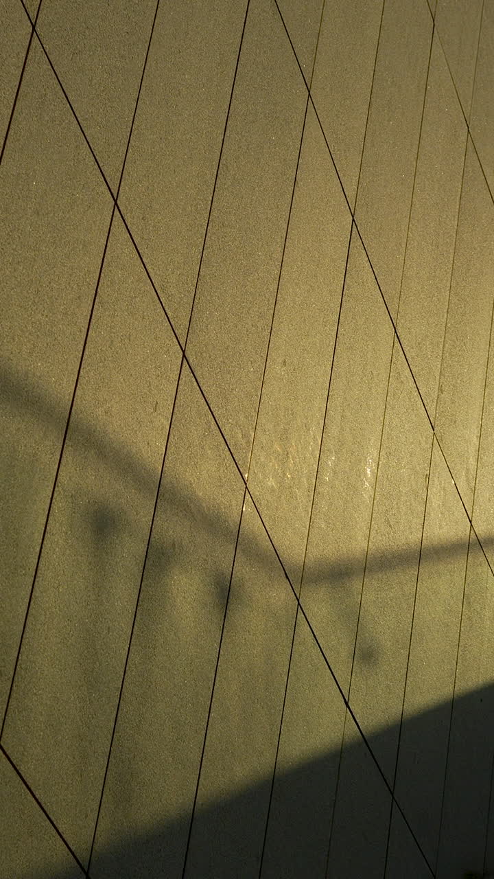 Imagem vertical de sombras da hora dourada projetadas sobre uma parede arquitetônica texturizada com um padrão de grade de losangos. Ideal para fundos artísticos ou conceitos de design urbano.