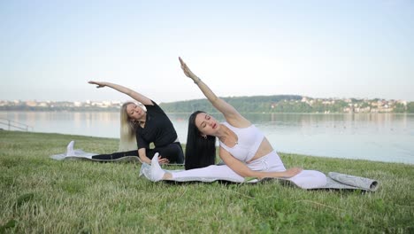 two beautiful slender young women are doing yoga she is sitting on a mat and raising her arms up two beautiful slender young women are doing yoga she is sitting on a mat and raising her hands up in the city park women practicing yoga outdoors on a sunny day
