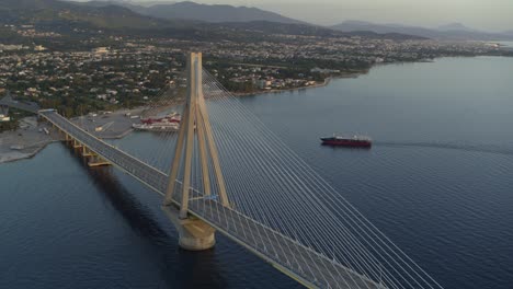 Luftaufnahme---Schiff,-Das-In-Der-Abenddämmerung-Hinter-Der-Brücke-Rio-Antirrio-In-Griechenland-Vorbeifährt---Aufnahme-Auf-Dji-Inspire-2-X-7-Raw