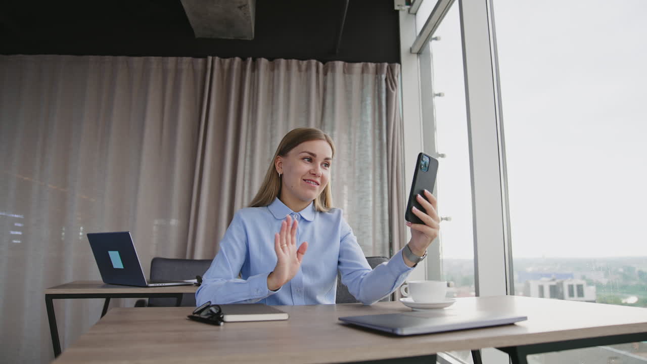 Good-looking lady in blue shirt looking on the phone in front of her. Smiling woman having conversation via online video chat. Cityscape at backdrop in window.