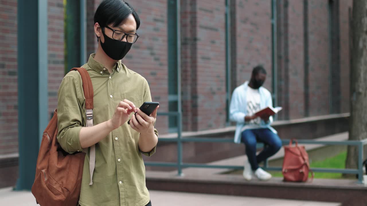 Camera zooming on asian student wearing glasses and facial mask using his smartphone near the college