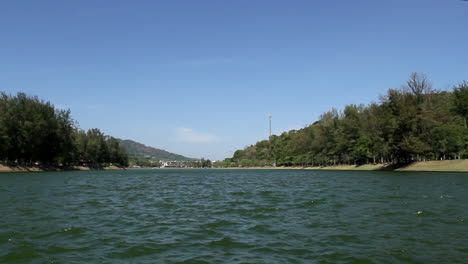 locked low angle shot of panoramic view of nai harn lake on a breezy blue sky summer day, no people, water in foreground and idyllic park and forest in background