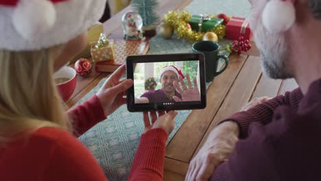 Smiling-caucasian-couple-with-santa-hats-using-tablet-for-christmas-video-call-with-man-on-screen