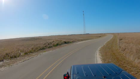 vehicle top pov while driving thru a curve on paved road thru grasslands and towards cell tower