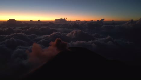 cinematic drone orbit captures fuego volcano smoking after eruption, clouds blanket valley at sunset, vibrant sky hues