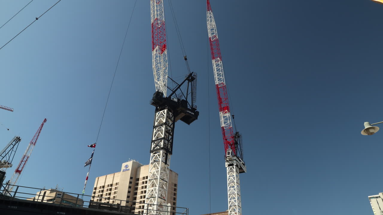 Two large red and white construction cranes stand tall against a bright blue sky. This tilt-down shot captures the scale and power of these engineering marvels.