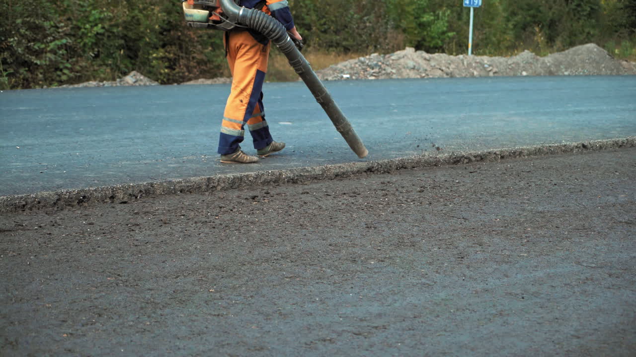 Worker in protective clothes blows out the pieces of old asphalt from the pits at road construction site. Road works concept. Worker is using compressed air to blow off dust on the road.