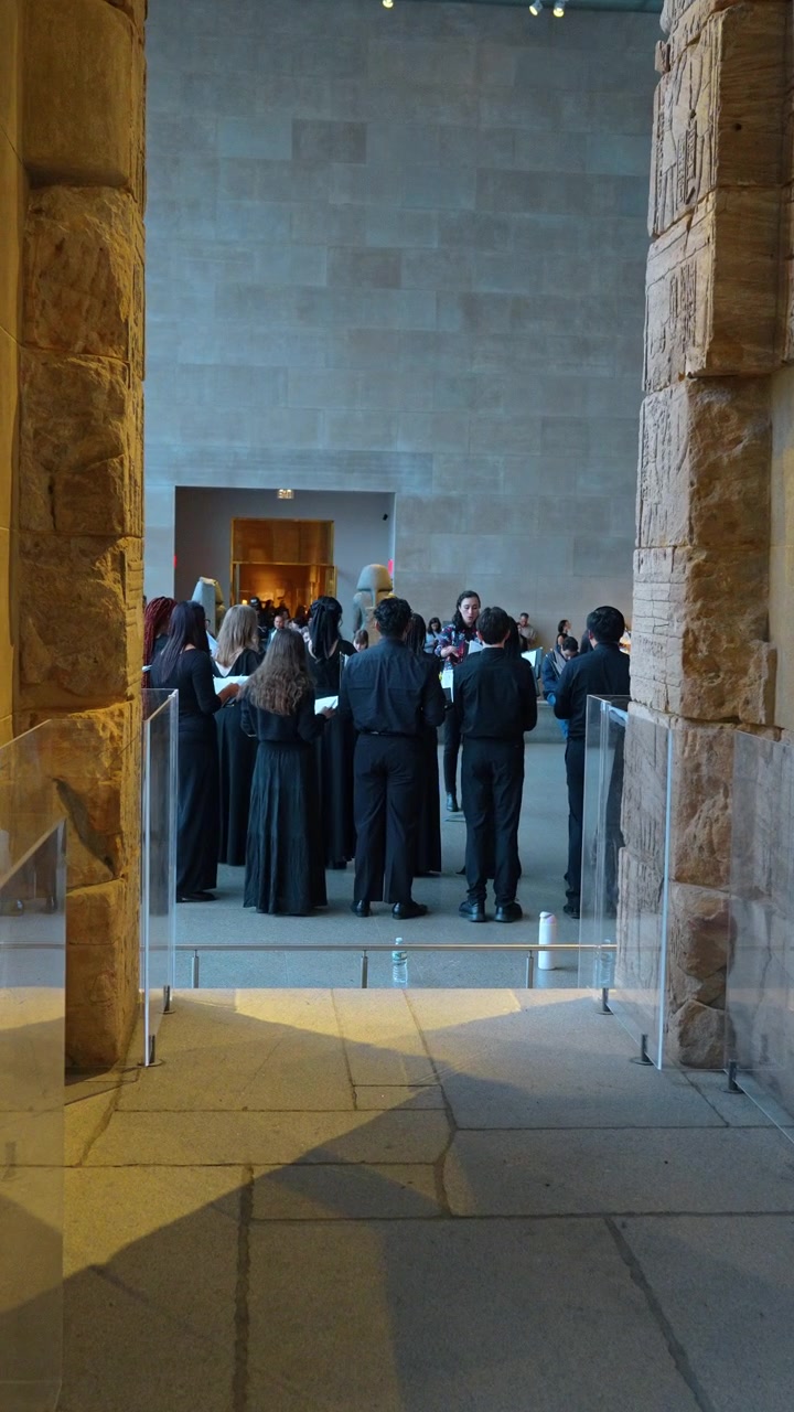 New York, USA, 25 maggio 2026: Esibizione di un coro incorniciata da antiche colonne di pietra in un museo di New York. Coristi in abiti neri in piedi all'interno della sala con una parete scolpita sullo sfondo.