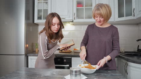 young girl attentively reads a cookbook while observing her mother stirring batter in a modern kitchen, she checks if the recipe is followed correctly