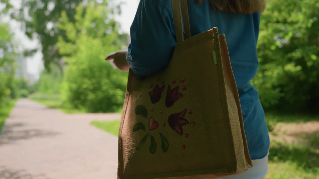 Back view of woman walking through lush green park carrying handbag, sunlight gently shining on long brown hair, peaceful summer atmosphere filled with warmth