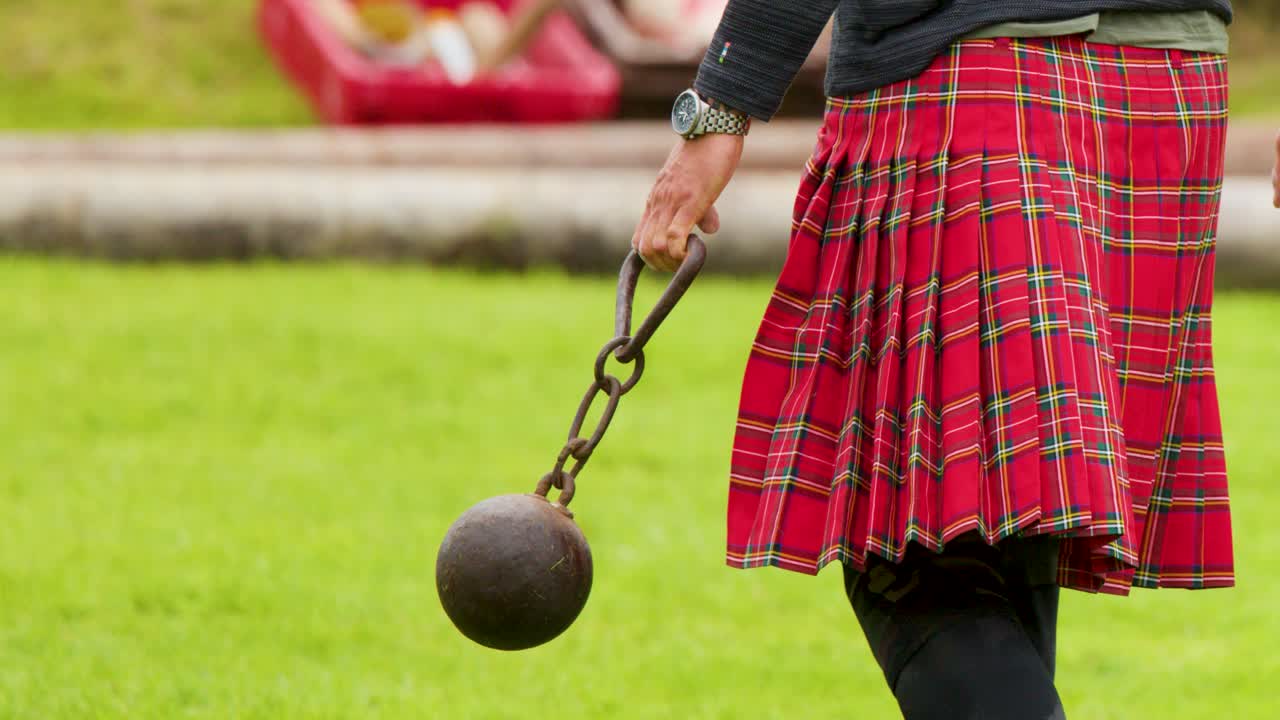 Man in tartan kilt walks on grass, carrying heavy hammer, outdoor daylight, mid-range shot