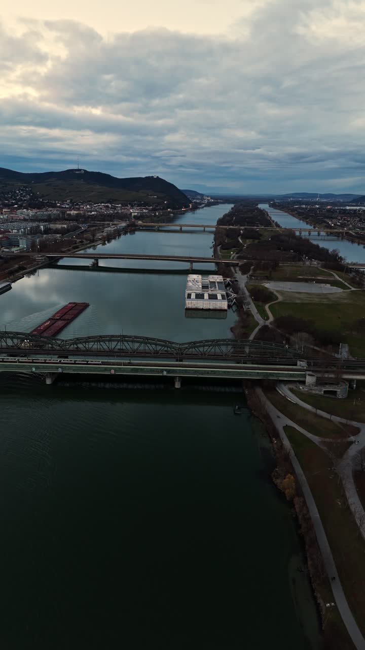 Aerial drone shot flying down the Danube River in Vienna, showing bridges spanning the water, boats moving along the river, surrounding city buildings, and cloudy winter skies overhead