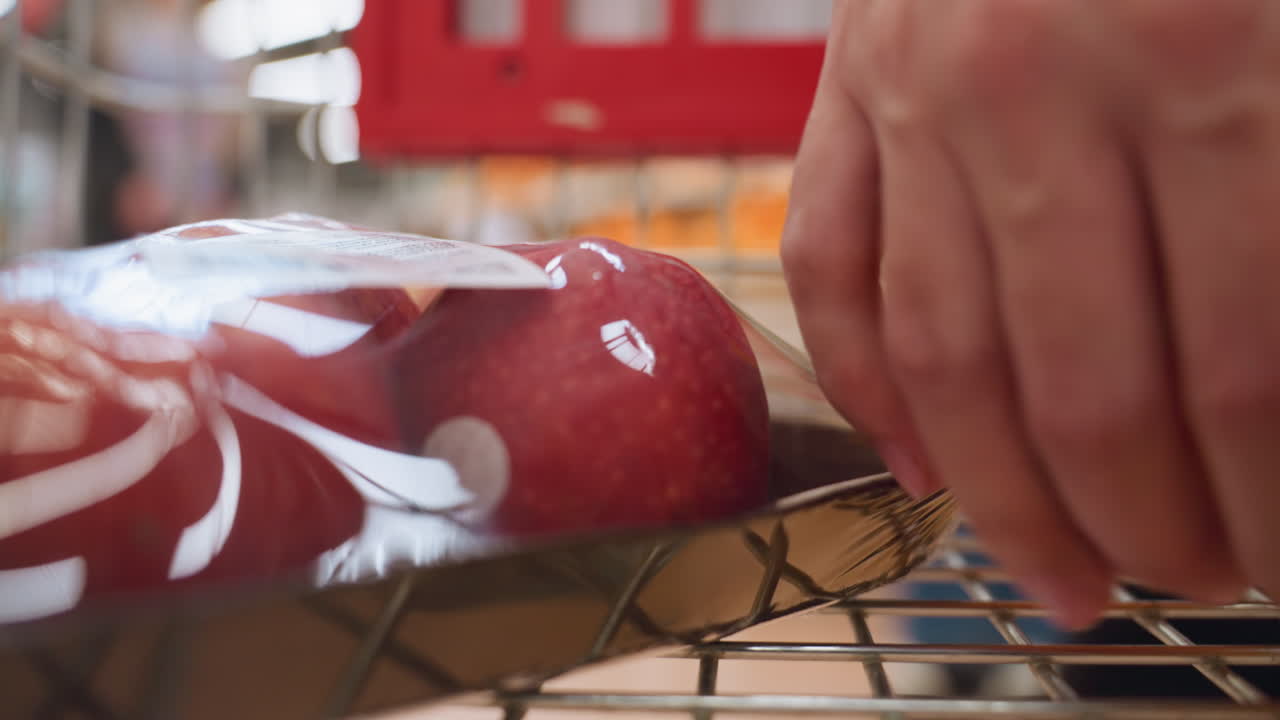 포장된 사과와 오렌지를 쇼핑 트롤리에 넣는 손. 이 장면은 식료품 쇼핑의 행위를 강조하며, 드 헬드 카메라로 촬영하여 역동적이고 몰입력있는 시야를 제공합니다.