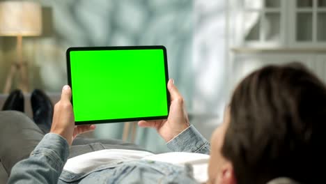 vista por encima del hombro joven en casa acostado en un sofá usando con pantalla de maqueta verde tableta computadora en modo horizontal. hombre usando dispositivo de pantalla táctil, navegando por internet