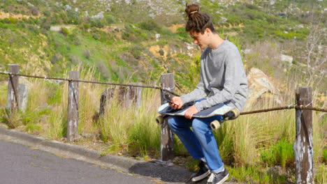 side view of cool young caucasian man with skateboard using mobile phone at countryside road 4k