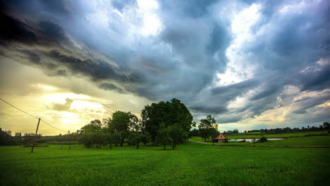 Kleiner-Bauernhof-Auf-Einer-Wiese,-Sich-Bewegende-Wolken-Im-Zeitraffer,-Stimmungsvoller-Himmel,-Gelber-Sonnenuntergang,-Großer-Grüner-Baum-Im-Grasland,-Kleiner-See-Im-Zeitraffer,-Aufnahme-Einer-Einrichtung,-Ländliches-Gebiet,-Ländliches-Ackerland