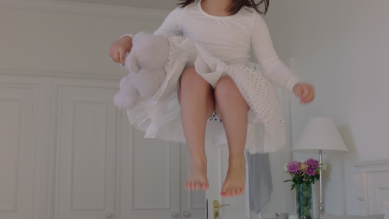 아름다운 작은 소녀가 침대에 뛰어 들고 테디 베어를 들고 재미있는 아이가 놀이 분위기에서 주말 아침을 집에서 즐기고 있습니다.