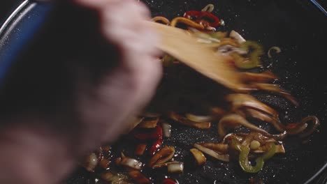 slices of onions and peppers in a frying pan, cooking and mixing them with a wooden spoon, close up view from above