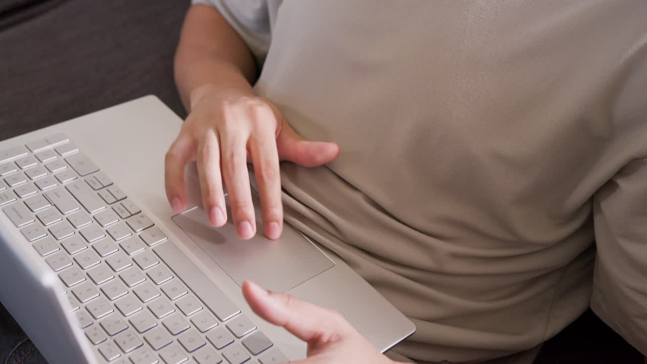 Adult man typing, tapping touchpad on sofa in tan shirt, black bracelet, working on silver laptop