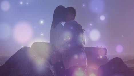 couple embracing and kissing on rocky coast, showing marketing graphics with pastel bokeh flares