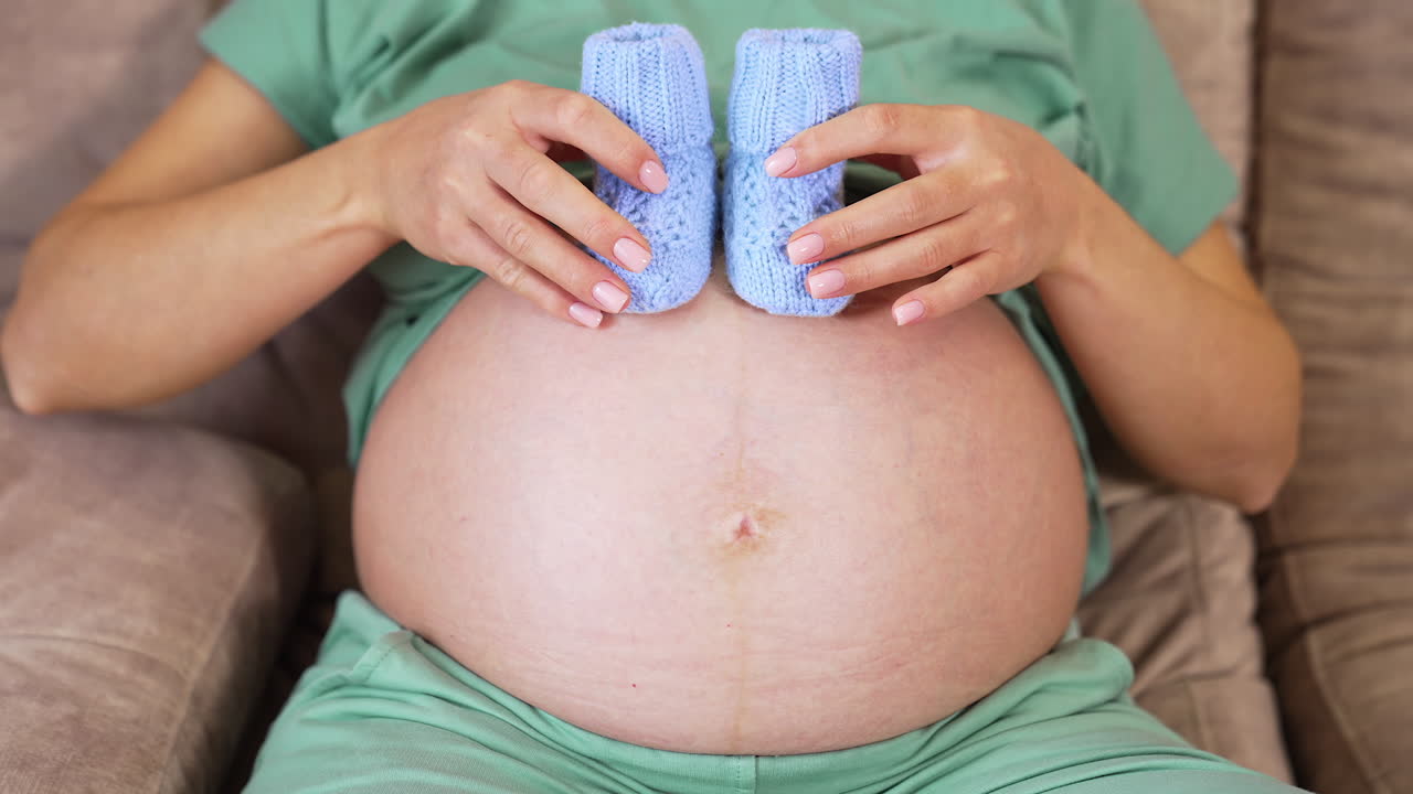 Big belly of pregnant woman where she puts tiny knitted socks. Future mommy shows heart sign with her hands. Beautiful little baby socks on the belly closeup.