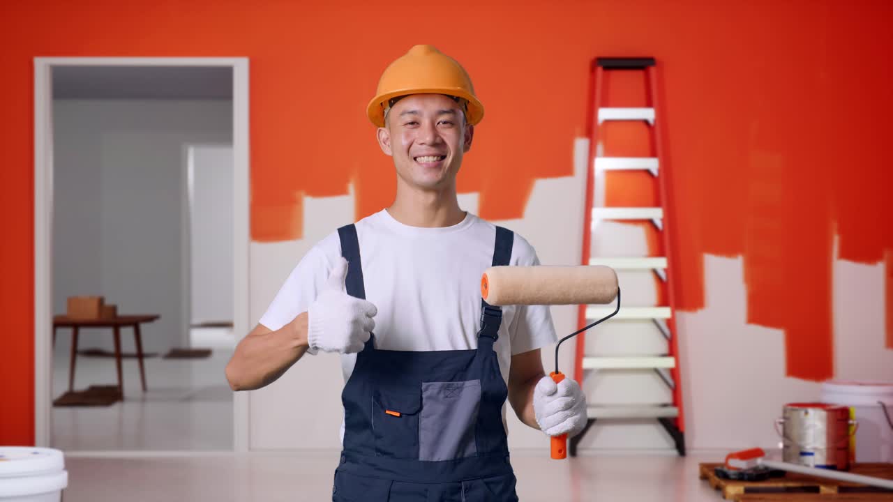 Asian Man Painter Wearing Safety Helmet Smiling And Showing Thumbs Up Gesture While Standing With Painting Wall In Room