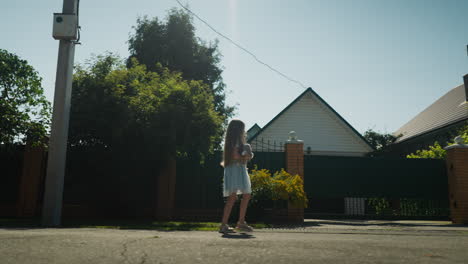 side view of young girl holding teddy walking past residential home as sunlight creates natural glow above her head, surrounded by greenery and fences on quiet street in warm afternoon light