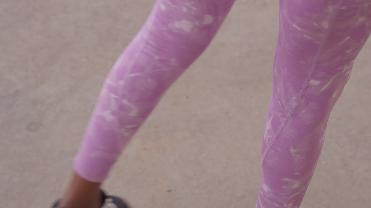 Close-up kaki wanita berkulit hitam bergerak dalam legging ungu selama jalan kaki pendinginan yang lambat dan penuh perhatian, skenario pendinginan yoga dan jalan kaki gaya hidup, langkah yang stabil, postur dan kesejajaran pergelangan kaki di atas beton halus.