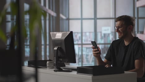 smiling young guy using mobile apps sit at table. male millennial professional holding modern smartphone texting message in office. young businessman using helpful mobile apps for business time