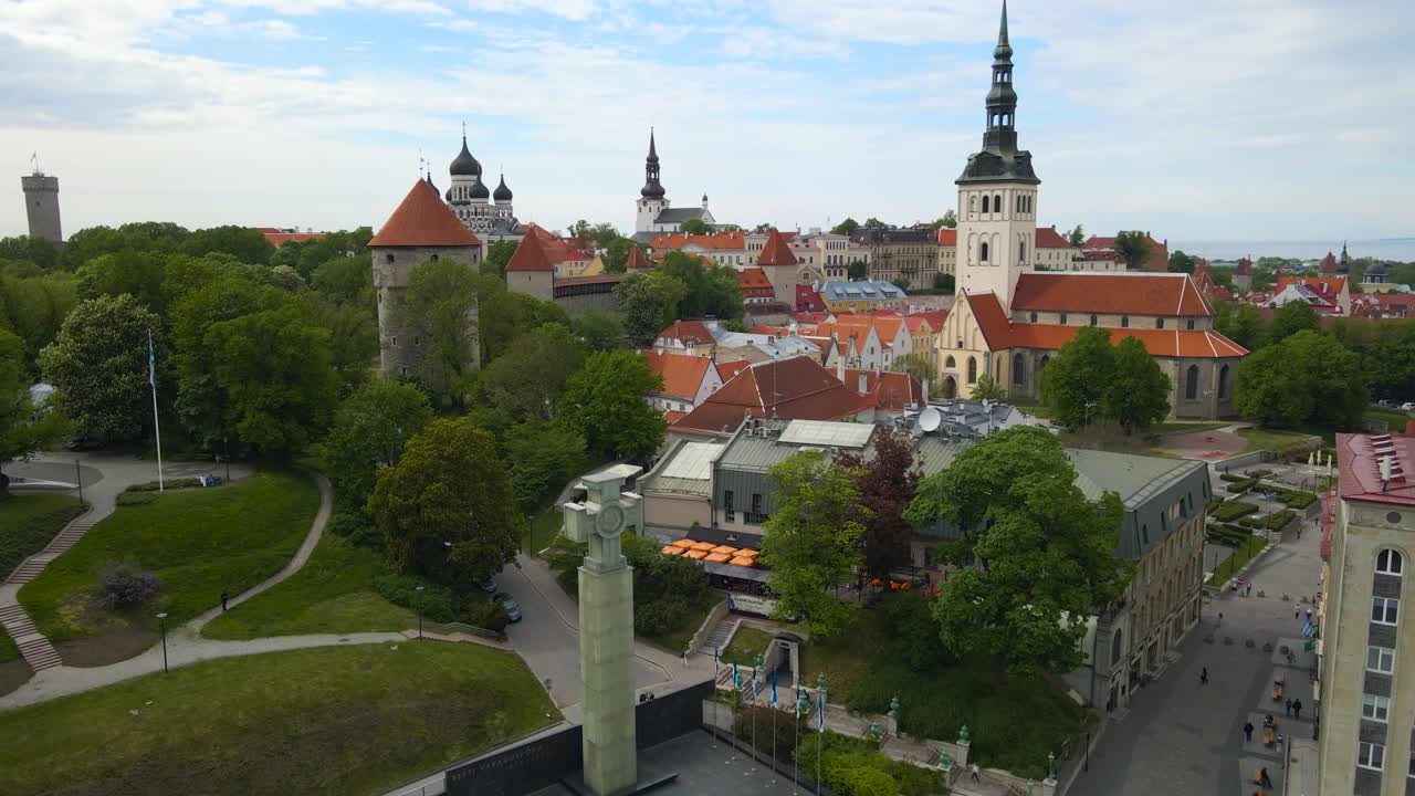 Aerial drone footage flying backwards and revealing Tallinn central city medieval buildings and a large victory cross monument in Freedom square during a summer day. Vurch towers and red rooftops.