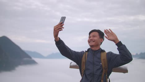 asian hiker male waving hand and speaking to smartphone while live stream the view of top foggy mountain