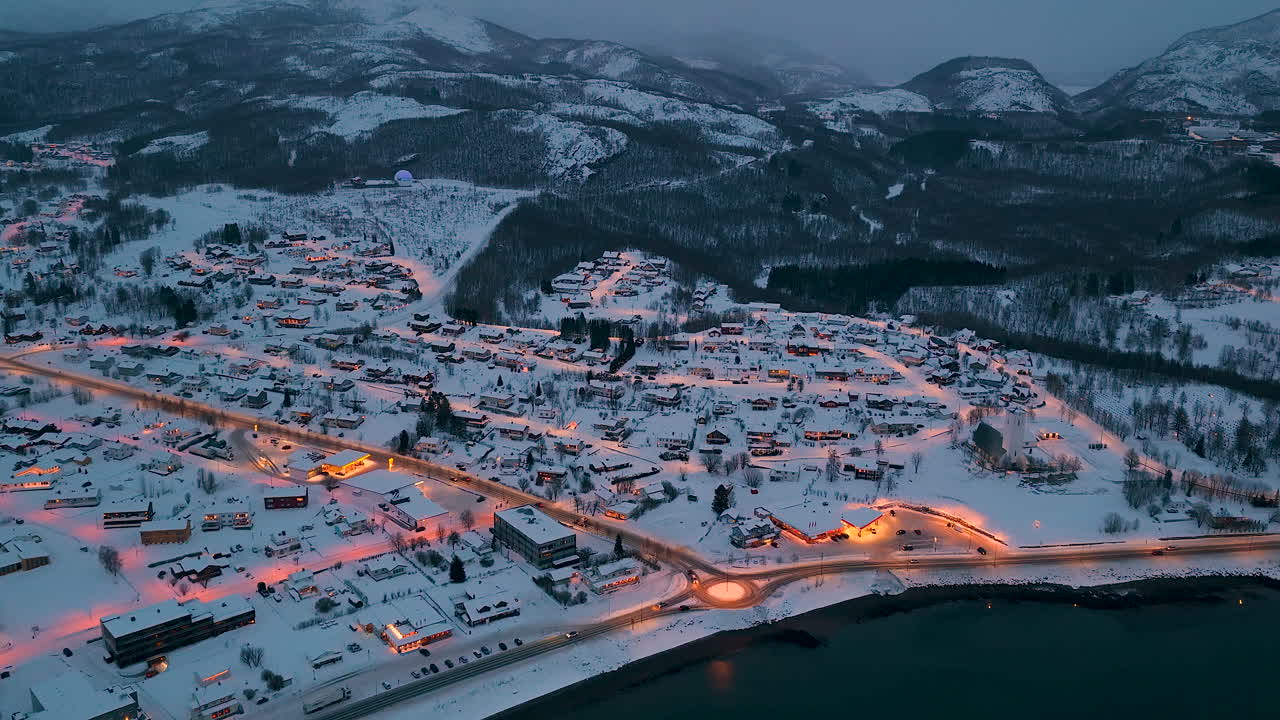 Korkealta otettu ilmakuva lumipeitteisestä norjalaisesta kylästä Lofootien saaristossa sinisen tunnin aikaan, jossa on valaistuja katuja ja taloja tummien vuorten taustalla.